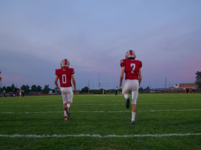 Several football players jog across the pitch in the evening light wearing matching red jerseys and helmets their movements framed by the warm glow of the setting sun