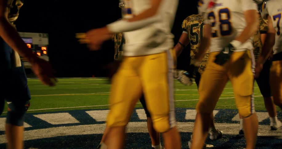 The video shows a football team on a field at night there are two distinct lines of players visible the players are walking and mingling in two distinct lines one in the foreground and one in the background the players in the foreground are wearin