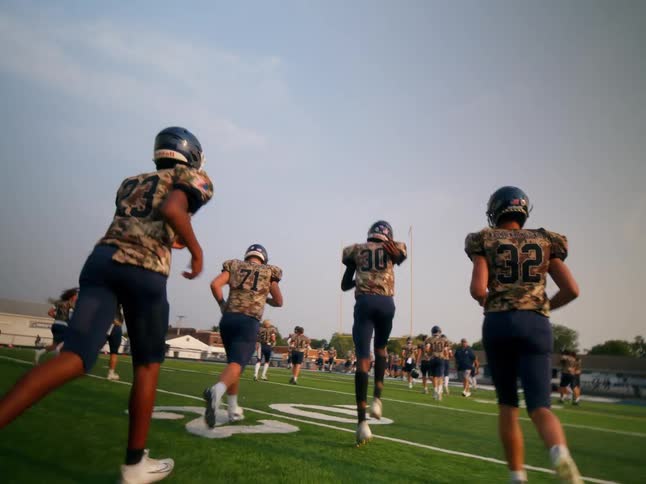 A team of football players in camouflage jerseys and dark shorts runs across a green field under a cloudy sky