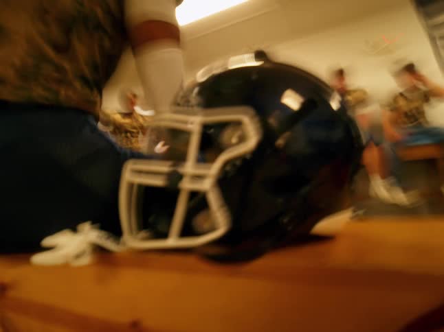 A close blurry shot focuses on a football helmet resting on a wooden bench with male athletes in uniform visible and out of focus in the background