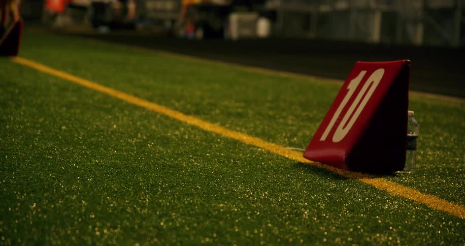 A football field with lush green grass featuring a bold yard marker sign displaying the number 10 positioned directly on a bright yellow line across the turf