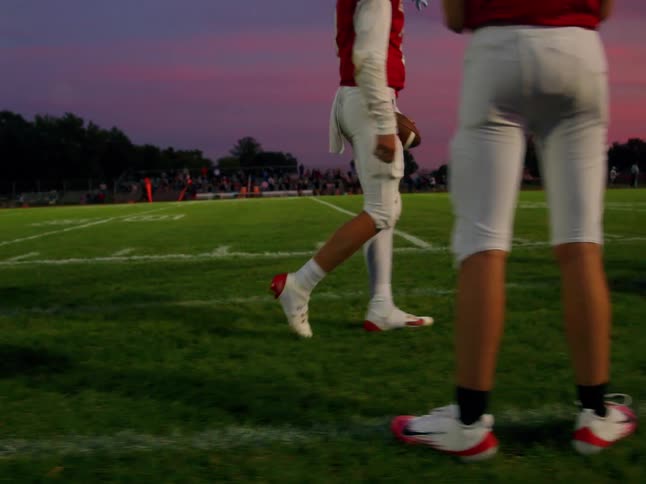 A young player walks across the field at night passing several football players as bright stadium lights bathe the scene in a dramatic glow creating a vivid electric atmosphere