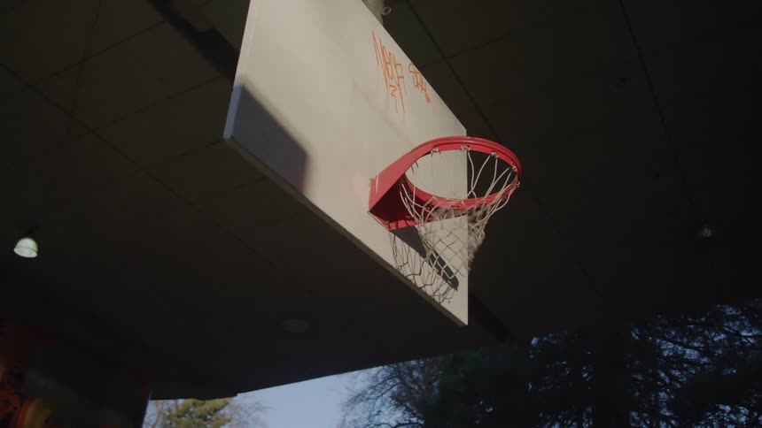 A lowangle view shows a basketball hoop and backboard attached to an overpass with graffiti on the backboard and the afternoon sunlight casting long shadows