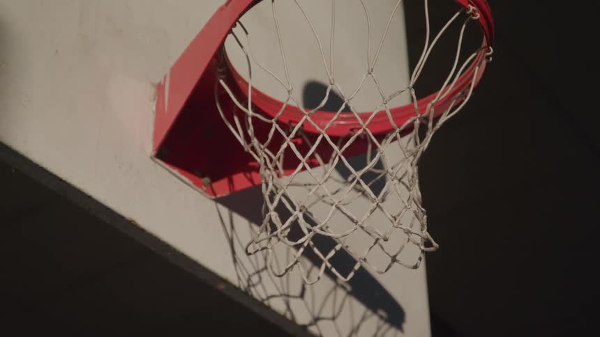 Based on the video a basketball flies into a hoop and passes through the net the hoop is red the net is white and the sun is casting a shadow behind it