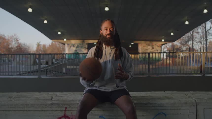 A man with a beard and dreadlocks is sitting and looking directly at the camera while holding a basketball under a bridge