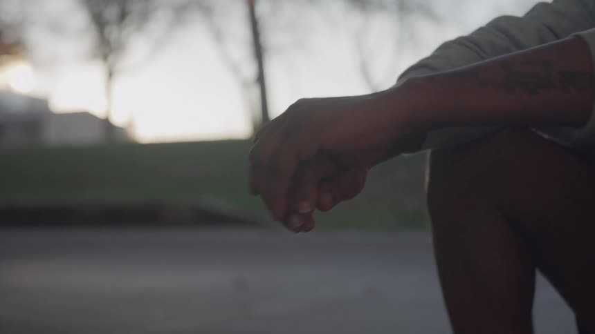 A close view of a mans arm and legs shows him sitting on the ground with his hands resting on his knees with tattoos visible on his forearm and a street scene with trees and buildings in the out of focus background during the evening light