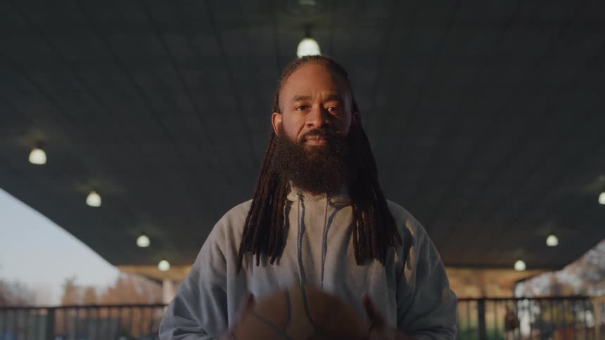 The video shows a man with dreadlocks and a beard holding a basketball he is wearing a grey hoodie and black shorts and is sitting under a bridge or overpass with lights in the ceiling the camera zooms in slightly as he looks directly at the viewer