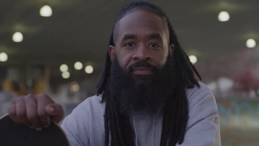 A person with a beard and long dreadlocks looks directly at the camera while holding a skateboard they are outside on what appears to be a basketball court