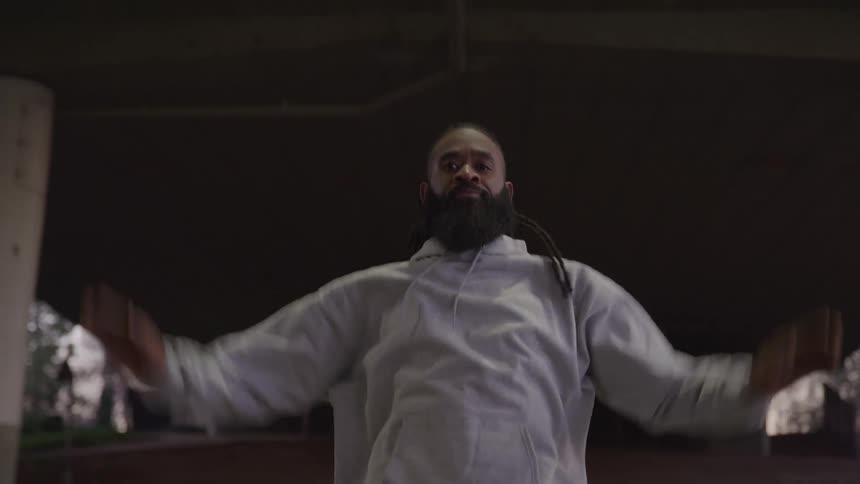 A bearded man in a white hoodie is doing jumping jacks or a warmup exercise under a concrete overpass