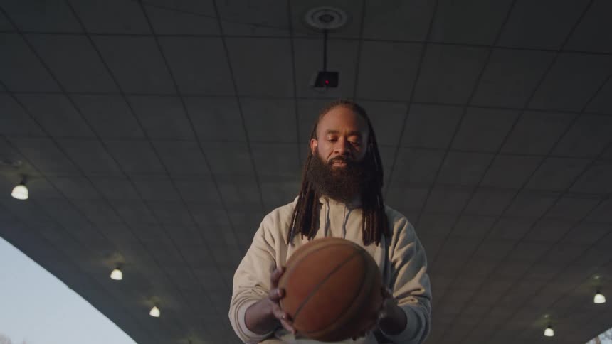 A male basketball player with long dreadlocks and a full beard sits on a concrete ledge with graffiti on it hes wearing a lightcolored sweatshirt and shorts while dribbling the basketball in his lap