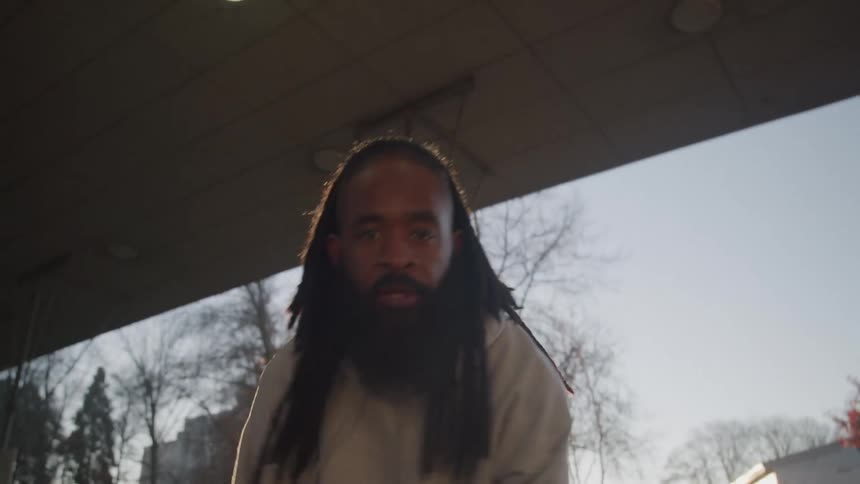 Based on the video a man with a beard and long dreadlocks is dribbling a basketball he is wearing a beige hoodie and red and white striped pants and is playing on an outdoor court under an overpass during either sunrise or sunset