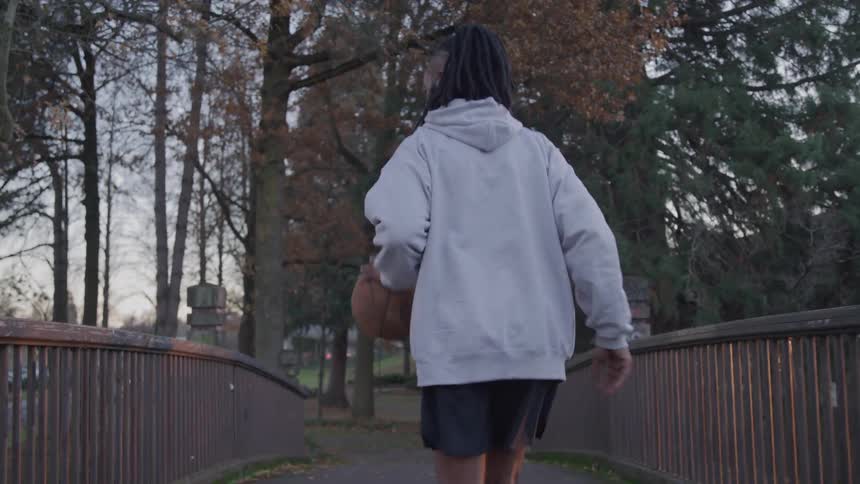 The video shows a person from behind wearing a grey hoodie and black shorts they are walking across a small bridge with a basketball tucked under their arm the path is lined with trees and the light suggests it is either early morning or late afternoo