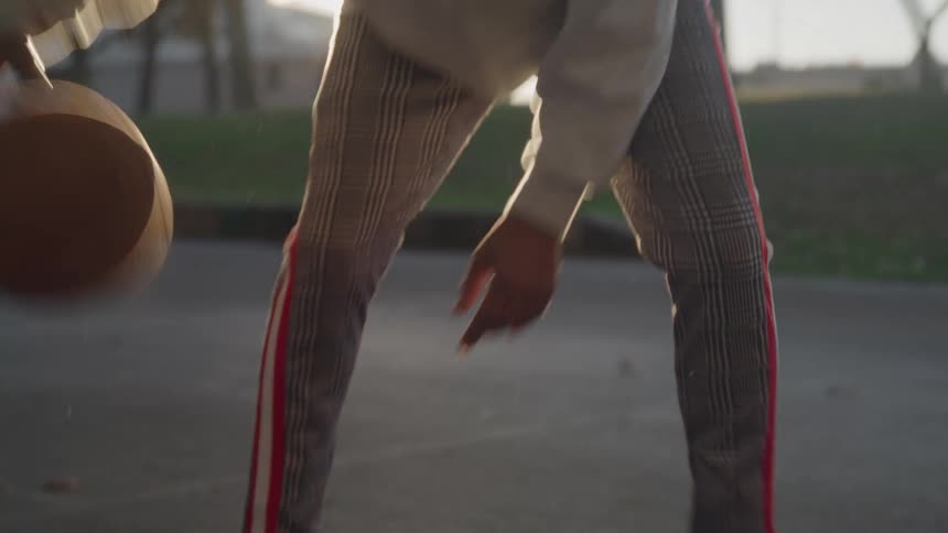Based on the video a man with long dreadlocks and a beard is dribbling a basketball on a court he is wearing a beige hoodie and red and white striped pants and the sun is shining brightly behind him