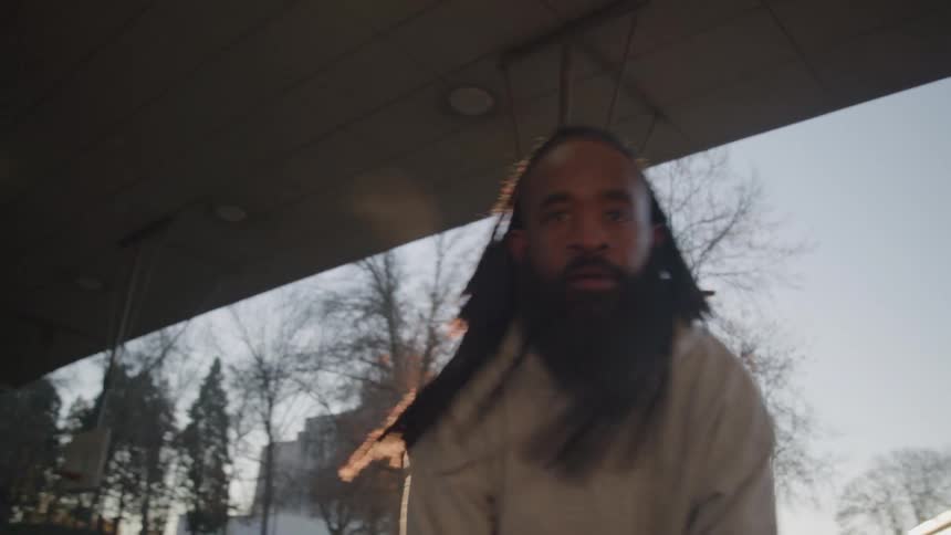 A man with dreadlocks and a beard dribbles a basketball on an outdoor court with sunlight flaring into the camera