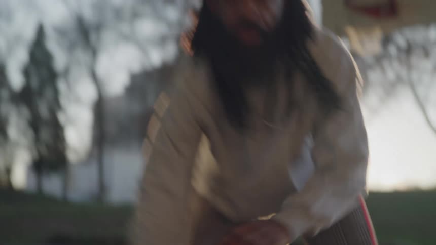 A male basketball player with long dreadlocks and a beard is shown dribbling a basketball on a street court at sunset wearing stylish gray plaid pants with red and white side stripes as the sunlight creates a bright lens flare