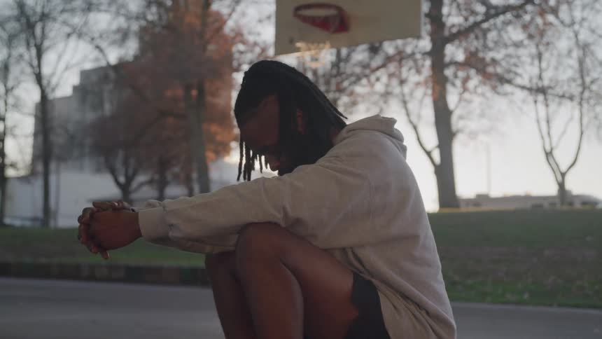 A black man sits on a basketball on an outdoor court