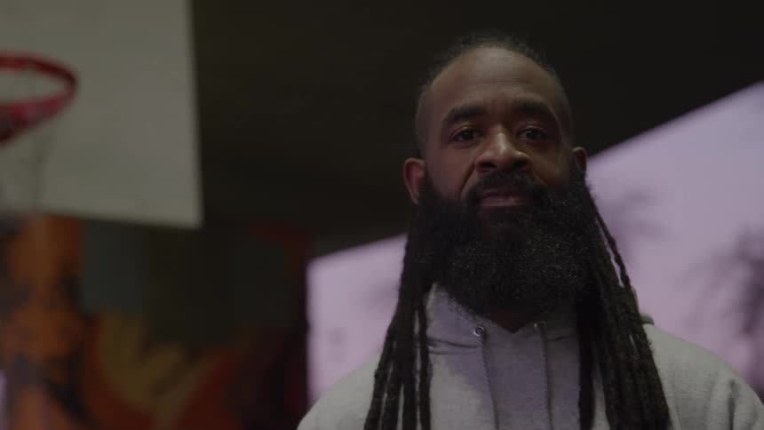Based on the video a man with a long thick beard and dreadlocks looks directly at the camera he is wearing a gray hoodie and standing in a gym with a basketball hoop visible in the background he is holding a basketball that is partially visible at the