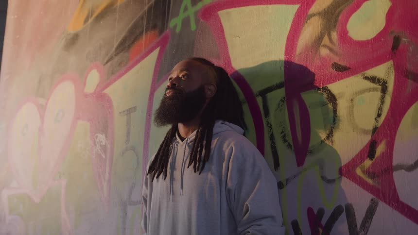 Man in white sweatshirt leaning thoughtfully against graffiti wall outdoors