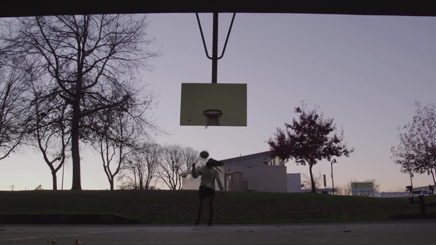Based on the video a young person wearing a hoodie and pants is dribbling a basketball they are outdoors on a basketball court at what appears to be either dusk or dawn the court is positioned in front of a small hill with some trees and a building in