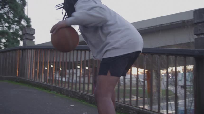 The shot follows a man with a beard and dreadlocks dressed in a grey hoodie and black shorts as he walks down a paved path he dribbles a basketball with skill as the sun sets in the background