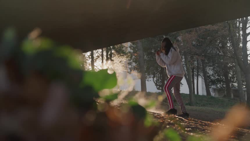 A male boxer in a gray hoodie and striped pants is warming up on a grassy field with morning sunlight creating a strong lens flare