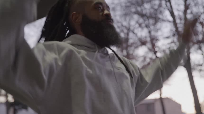 A man with dreadlocks and a long beard raising his arms in a calisthenics or warmup movement with the background of trees and suburban buildings slightly out of focus