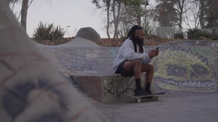 A black man with dreadlocks and a beard sitting on his skateboard in a concrete skate park and looking at his phone with graffiti on the walls behind him and tall trees in the background under a dim cloudy sky