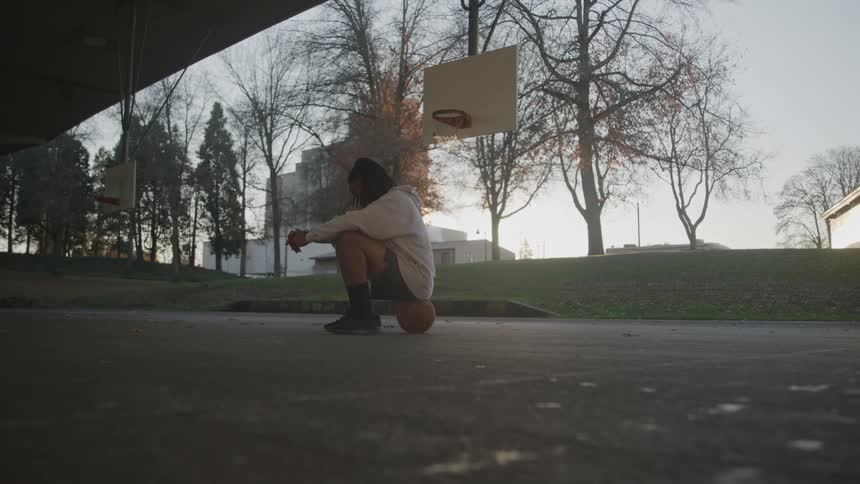 A lowangle shot shows a man dressed in a white hoodie sitting on a basketball court with his head down he is resting his hands on a basketball between his knees with a couple of basketball hoops in the background