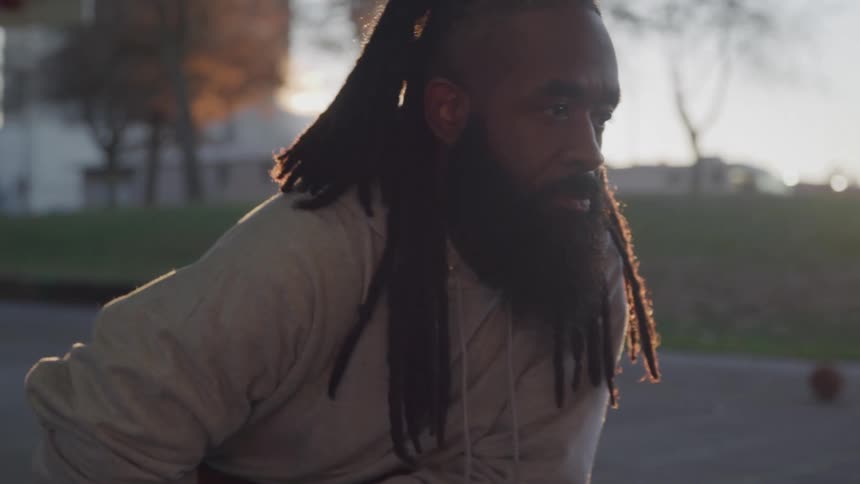 The video shows a man with dreadlocks and a full beard wearing a white sweatshirt and black shorts he is standing outdoors and appears to be in an athletic stance possibly stretching or preparing for a workout the sun is setting behind him creating a