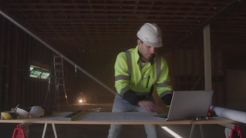 Male construction worker wearing a helmet and typing on a laptop computer on top of a table on a framed house indoors during day time