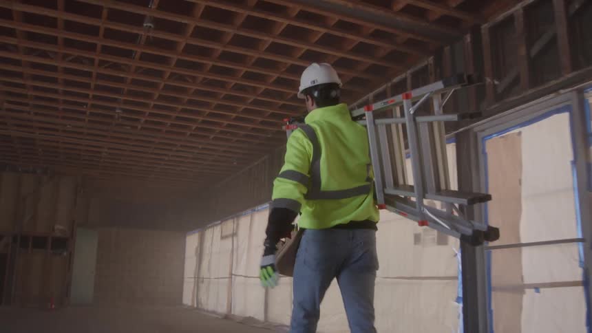 A male construction worker wearing a hard hat and reflective vest walks away from the camera in a dusty unfinished building while carrying a stepladder on his shoulder