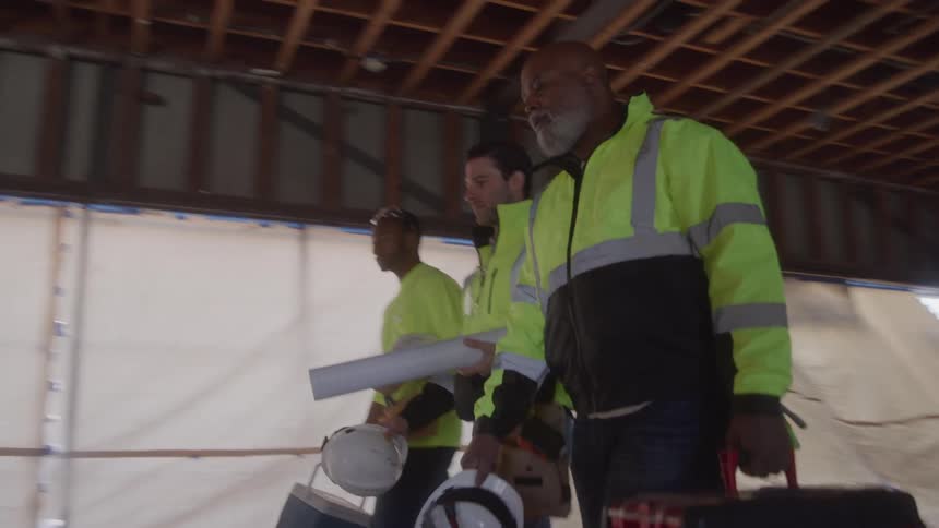Three construction workers walk through a dusty unfinished room carrying supplies they are all wearing hard hats and neon yellow safety vests