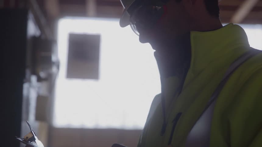 A close view of a male construction worker wearing a white hard hat and yellow reflective vest with sunlight from a nearby window silhouetting his body as he looks down at a clipboard