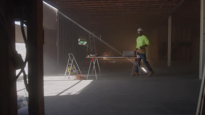 A person wearing a neon yellow longsleeved shirt and hard hat walks toward a workbench in a dusty room they stand behind the bench which has a laptop and appear to begin working on something