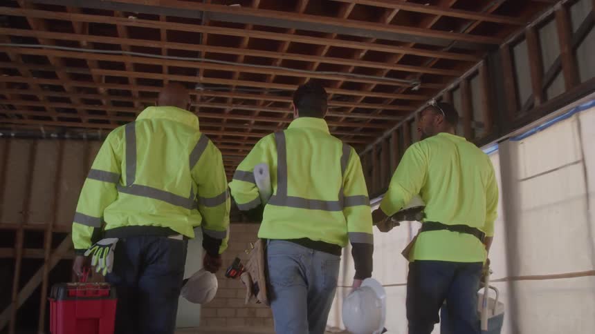 Three male construction workers in bright yellow vests and hard hats walk away from the camera in a building under construction carrying toolboxes and clipboards