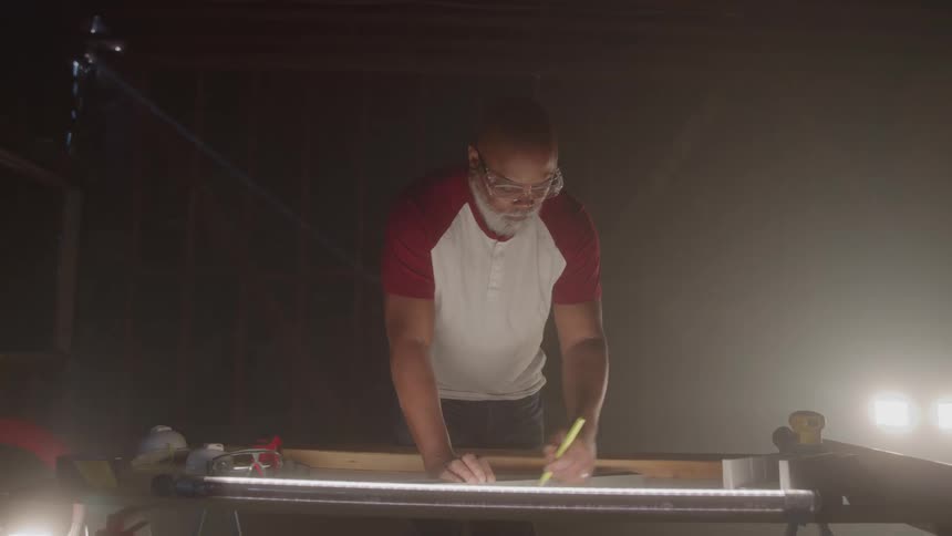 A male construction worker wearing safety glasses and a twotone shirt works at a workbench he leans over the bench and uses a pencil and a ruler to draw a line on a piece of paper
