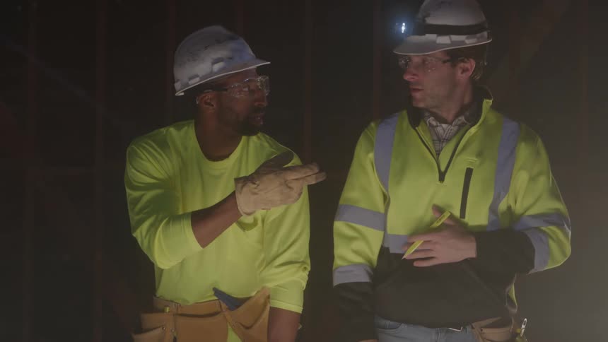 Two male construction workers wearing hard hats and reflective vests are talking in a dark unfinished building