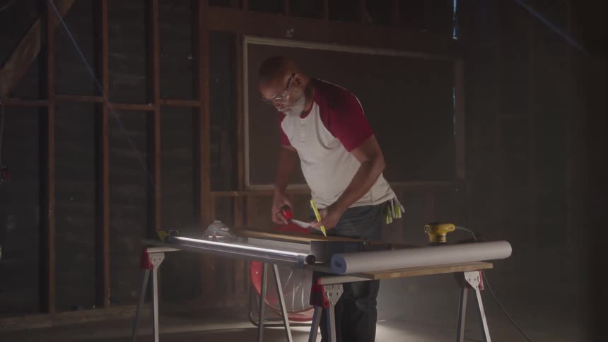A construction worker in safety glasses and a twotoned shirt uses a tape measure and a pencil he is leaning over a piece of wood on a workbench carefully marking a measurement