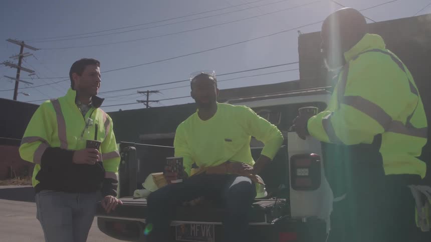 Three male construction workers in bright yellow vests are talking and drinking coffee while standing around the back of a pickup truck in a sunny industrial area
