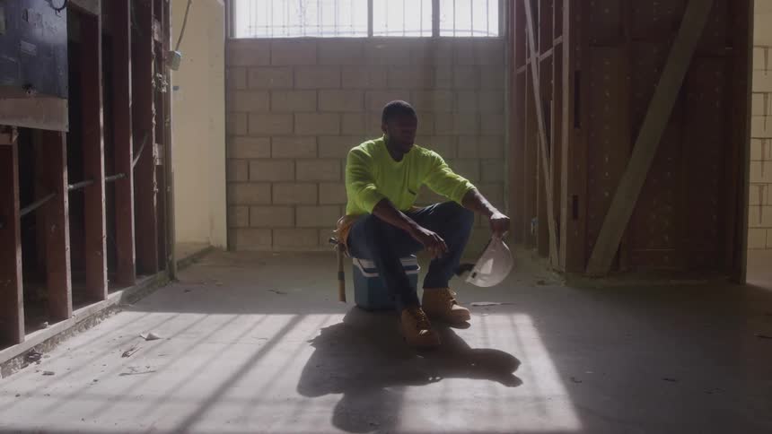 A tired male construction worker in a yellow shirt and boots sits on a blue cooler in a strippeddown dusty room taking off his hard hat with sunlight from a window behind him creating long shadows on the floor