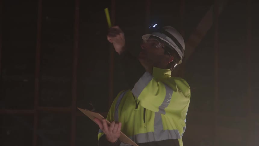 Based on the video a man is dressed in construction gear including a hard hat with a headlamp a safety vest and glasses he is holding a clipboard and a marker and appears to be inspecting something in a dimly lit unfinished building he uses the hea