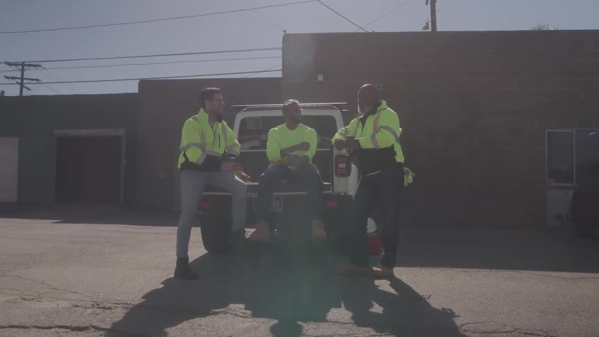 Three workers in reflective vests talk while leaning on the back of a vehicle