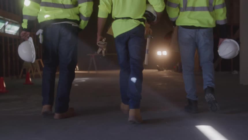 Three men wearing reflective yellow jackets and jeans walk away from the camera in a dark dusty warehouse carrying their hard hats and walking toward the light from an open garage door