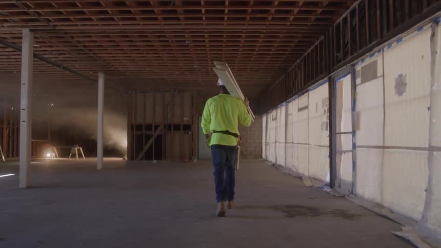 A male construction worker stands in a dark dusty room hes wearing a hard hat with a headlamp a bright yellow safety vest and glasses while holding a clipboard he looks up and to the side appearing to inspect the work site