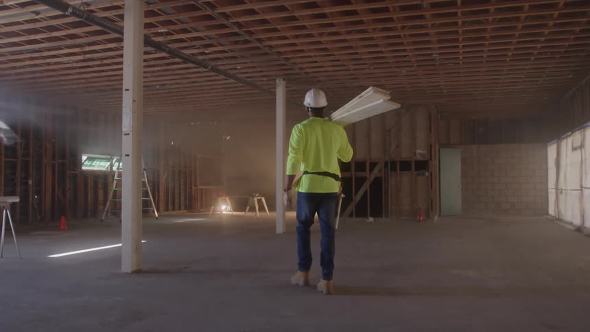 Worker wearing a helmet carries a drywall sheet on his shoulder inside a building under construction
