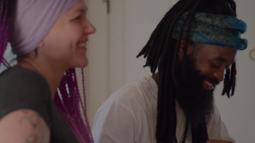 A smiling woman with purple braids and a man with dreadlocks and a blue headwrap share a cheerful moment together