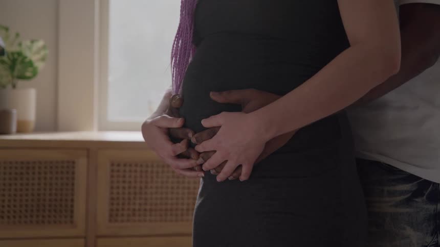 A man and a pregnant woman are standing close with the mans arms around the womans waist and the couples hands on her belly