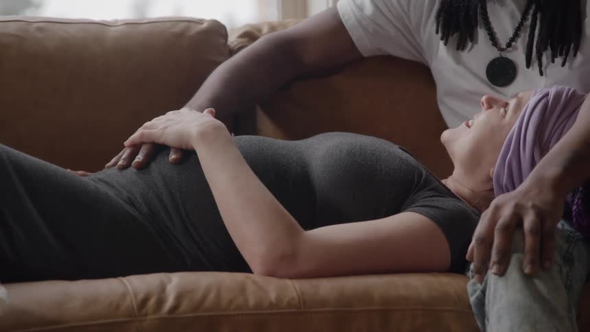 A young couple on a sofa the woman is lying and the man is sitting and both are with hands on her pregnant belly