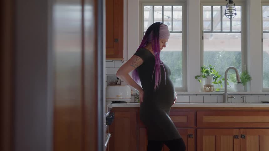 A pregnant woman with a head wrap is standing in her kitchen holding her baby bump and looking down at it with plants on the countertop and a window in the background