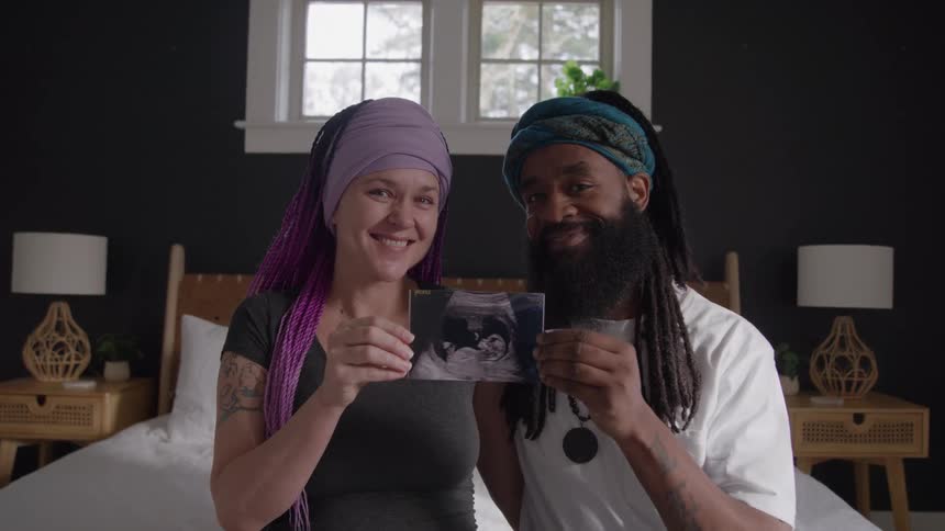 A man and a woman are sitting on a bed holding up a sonogram of a baby and smiling at the camera